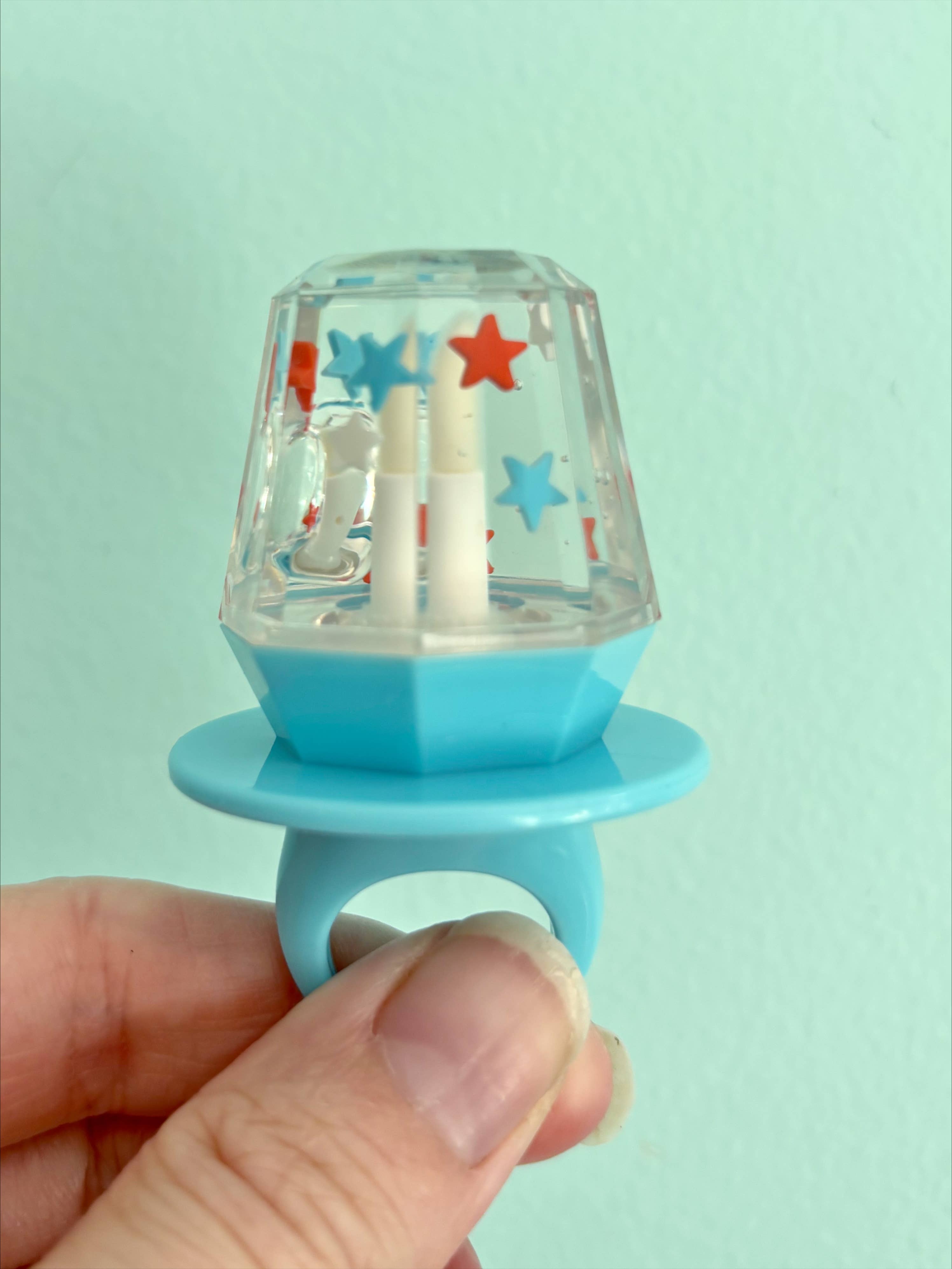 Blue lip gloss with a clear top featuring colorful star designs, held against a light green background.