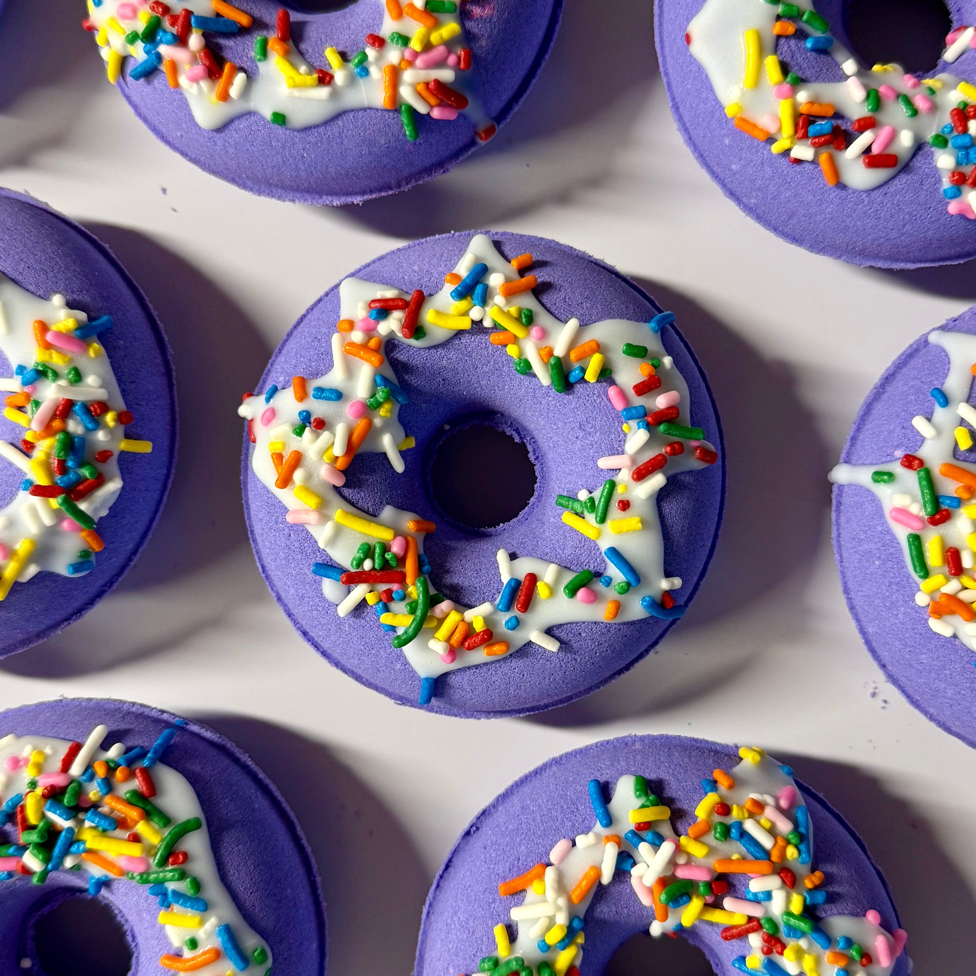 Purple donut bath bombs with white icing and colorful sprinkles on a white surface