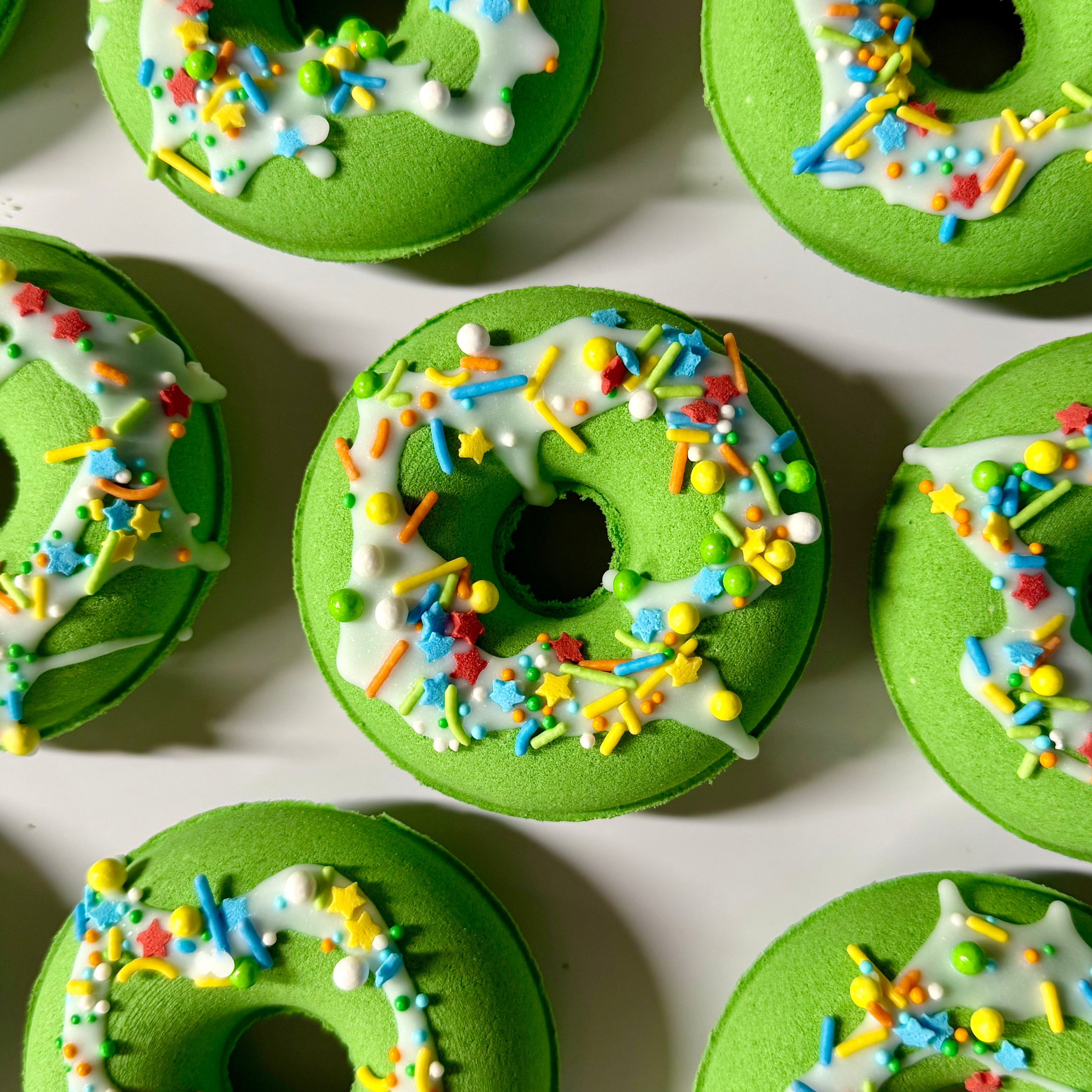 Green donut bath bombs with white icing and colorful sprinkles arranged on a light surface.